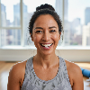 A natural, close-up portrait of a fit woman in her 40s, diverse ethnicity, with her hair tied back loosely. She has a post-workout glow and a big, genuine, open-mouthed smile, looking directly into the lens. She is wearing modern athletic wear (a tank top). The background is a bright, modern living room with large windows, a yoga mat, and a pair of dumbbells visible on the floor. Soft, natural window light. Shallow depth of field. Authentic lifestyle photography.